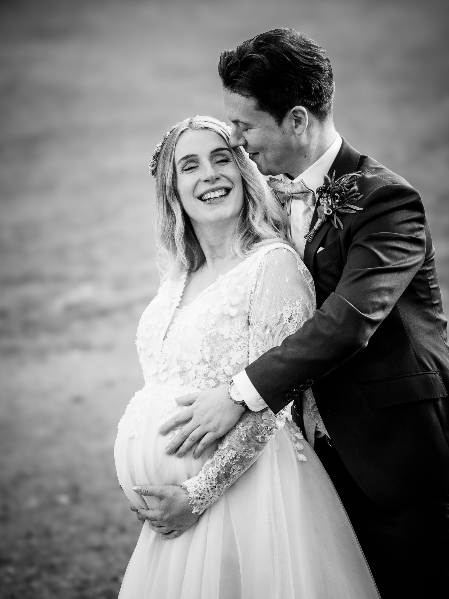 Pregnant bride smiles as groom embraces her baby bump in black and white wedding photo.