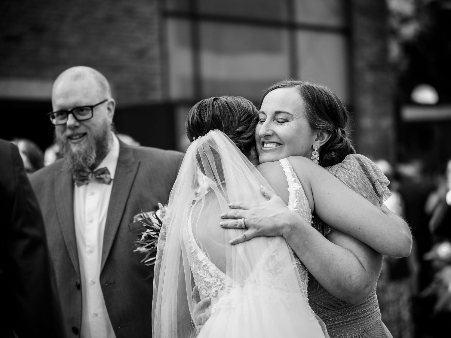 Black and white photo of a bride in a veil embracing a smiling woman at a wedding ceremony outdoors.