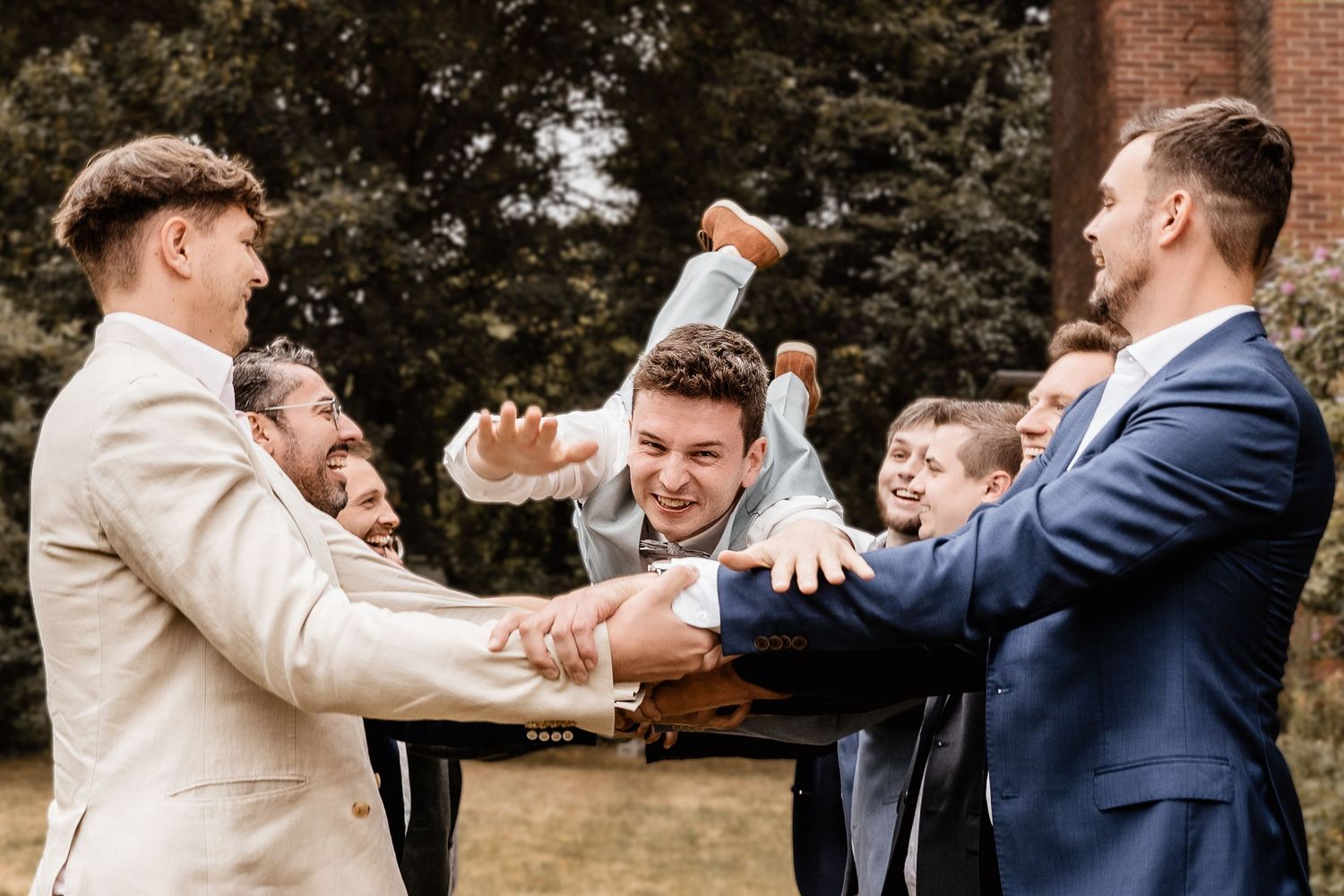 Groomsmen tossing groom into the air laughing outdoors in suits during wedding celebration.