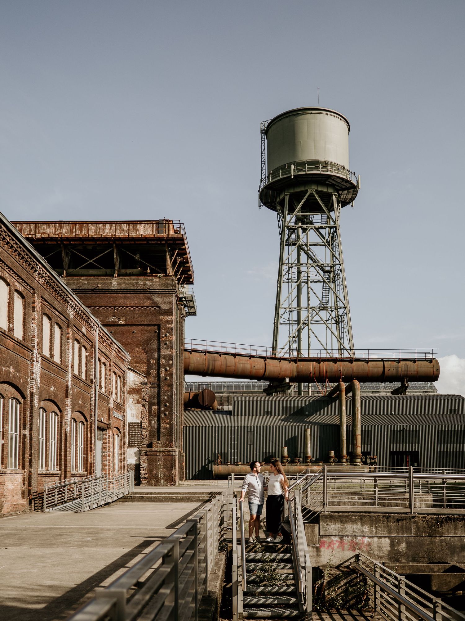 Couple walking on stairs at an industrial location with a vintage water tower and brick buildings backdrop.