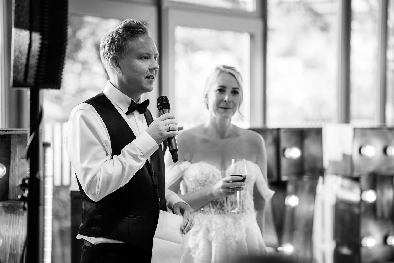 Groom in bow tie speaking into microphone beside smiling bride during wedding reception speech.