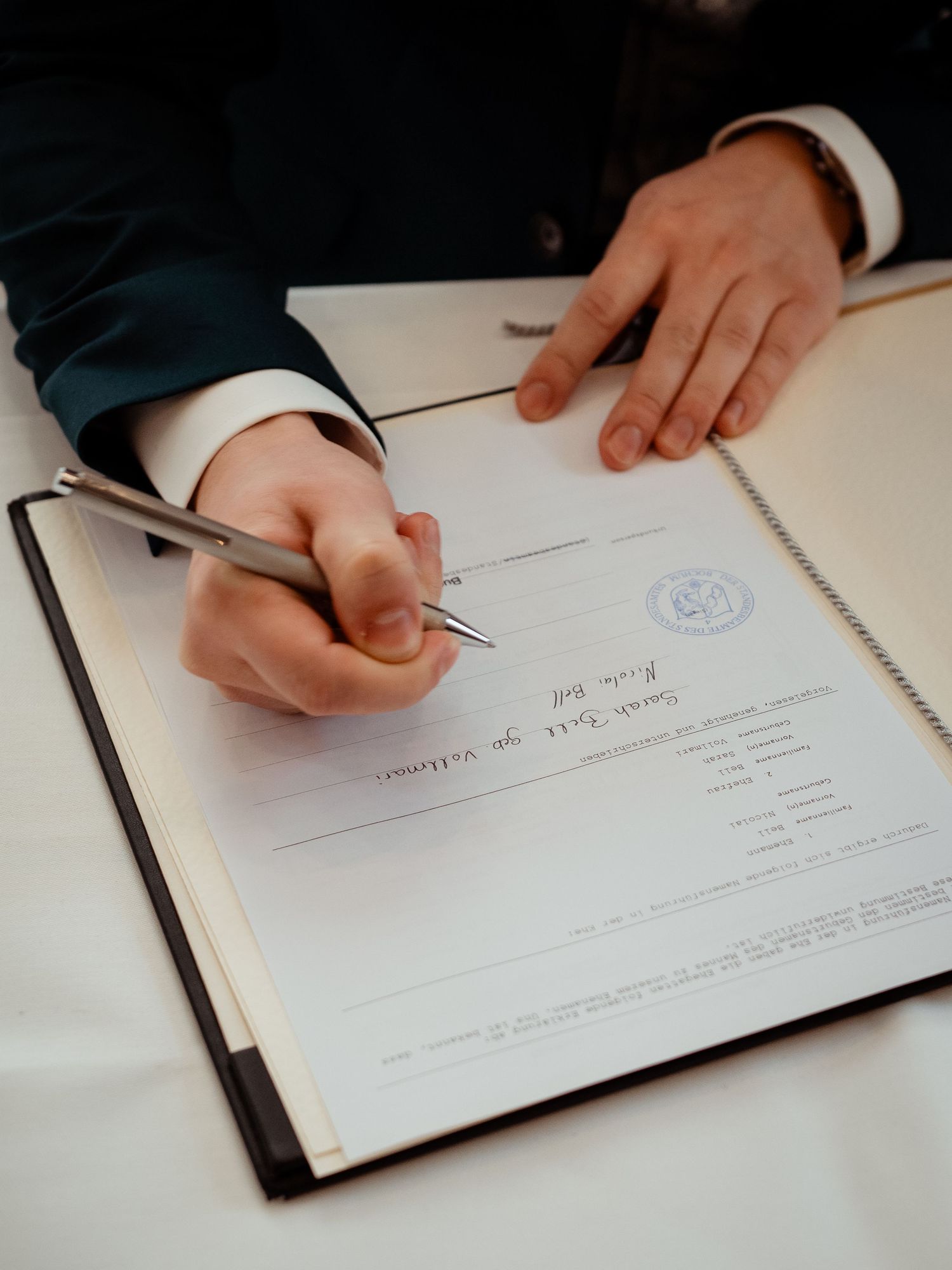 Groom in navy suit signs official wedding certificate document with silver pen at ceremony.