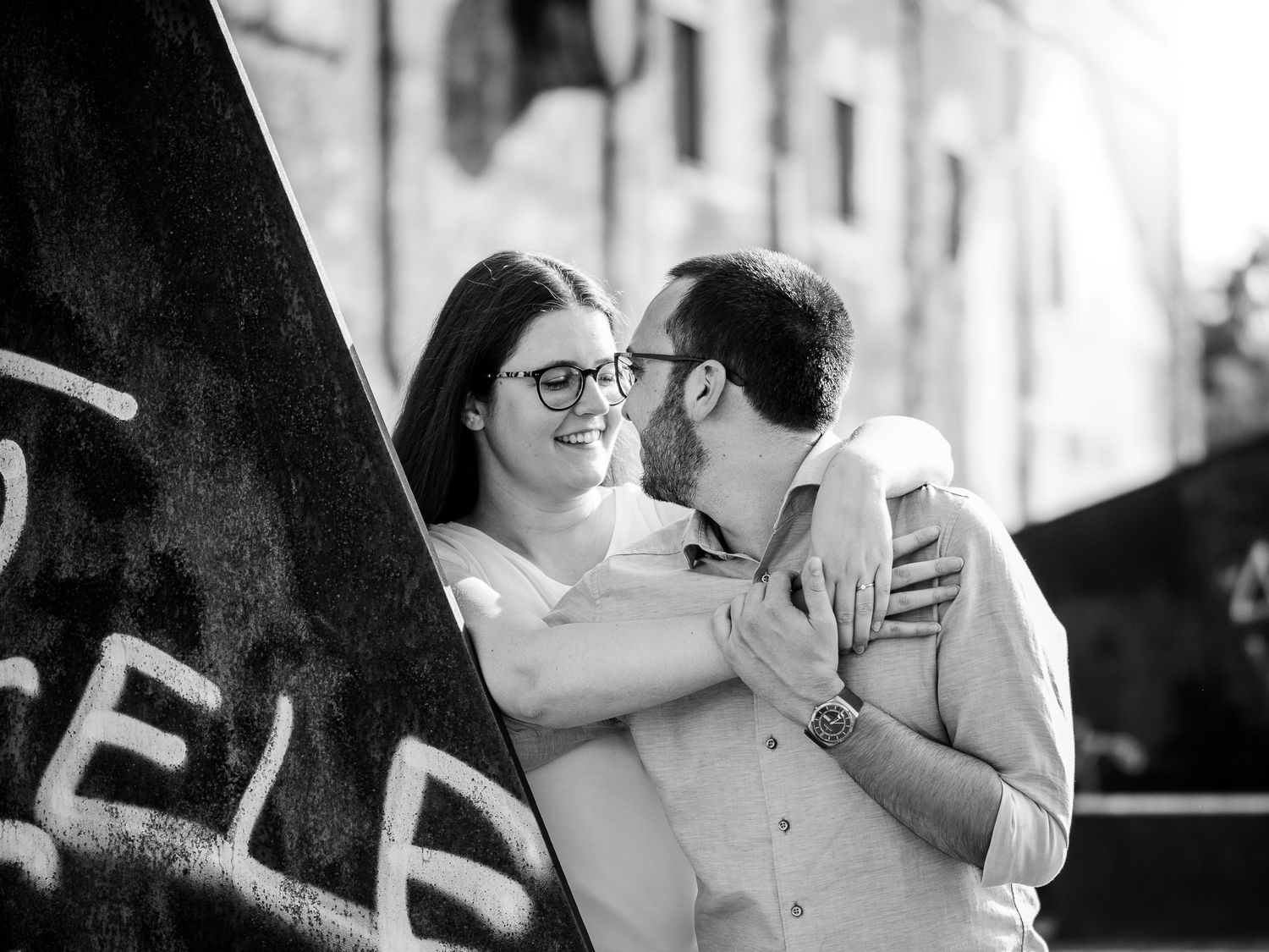 Black and white photo of a couple embracing and smiling near a graffiti wall outdoors.