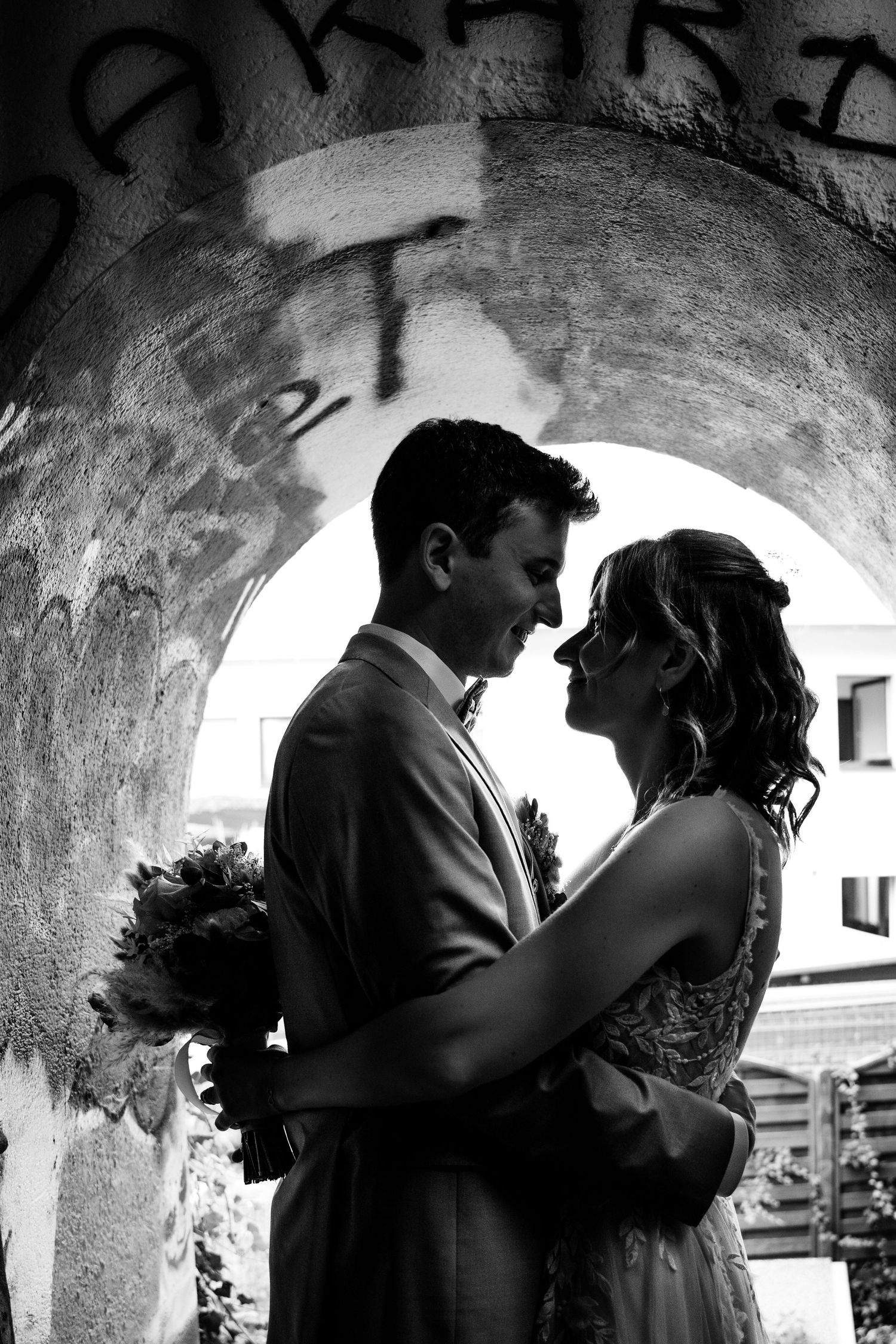 Bride and groom embrace romantically under a stone arch in a black and white wedding portrait.
