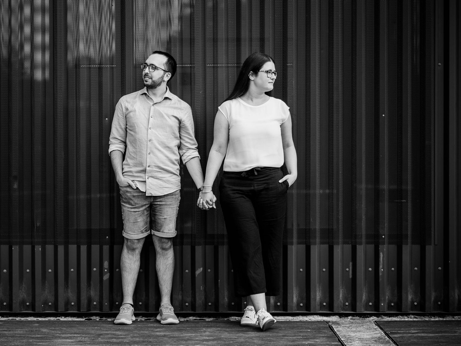 A black and white portrait of a couple holding hands while standing against a striped wall.