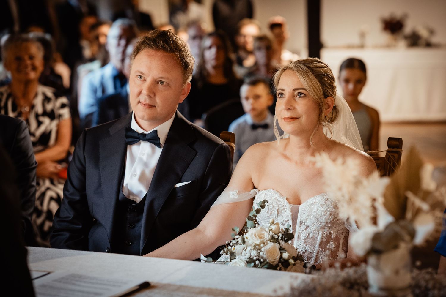 Bride and groom sitting together at wedding ceremony altar, bride in lace gown with veil, groom in black tuxedo.