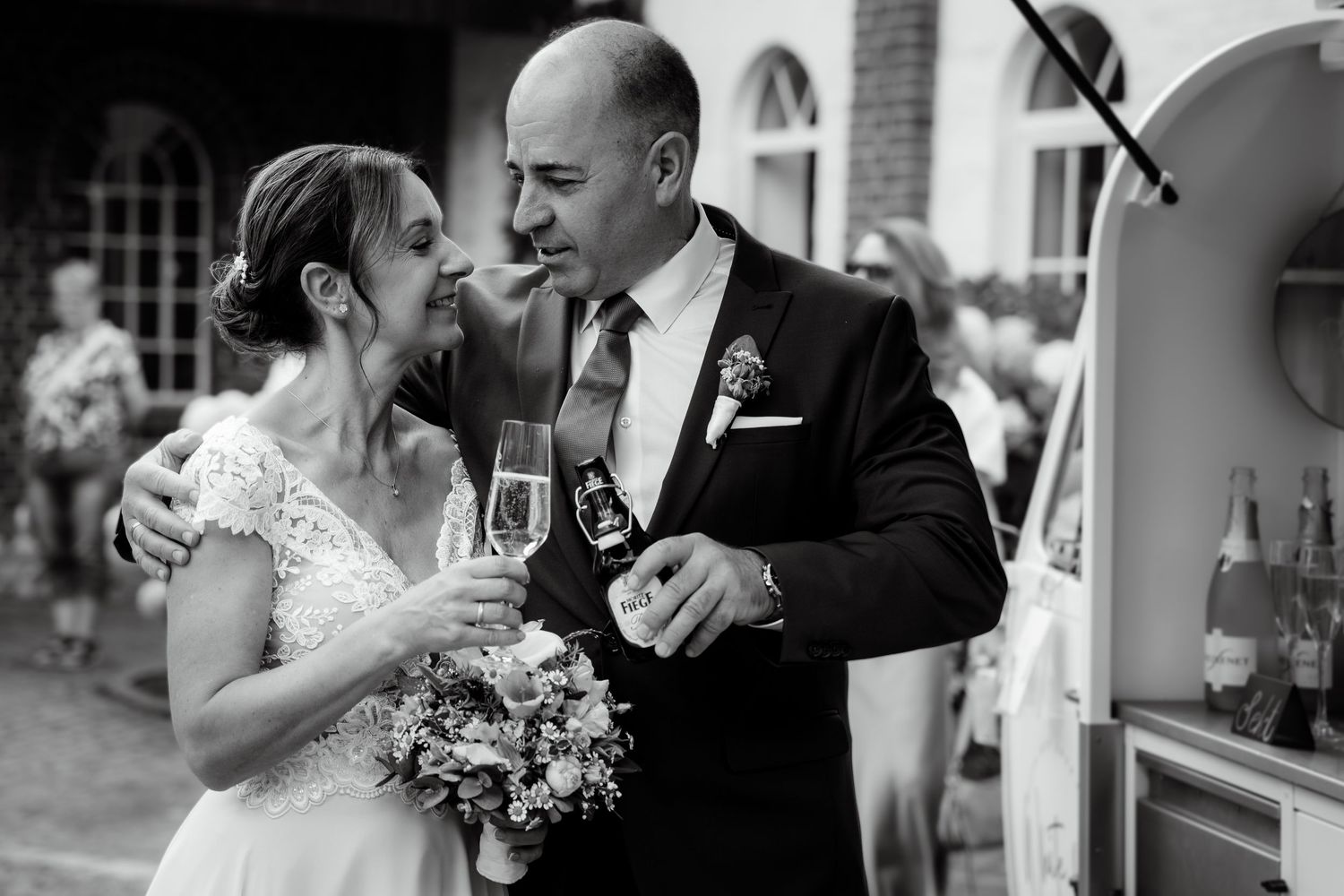 Bride and groom share champagne toast with bouquet in black and white candid wedding reception photo.