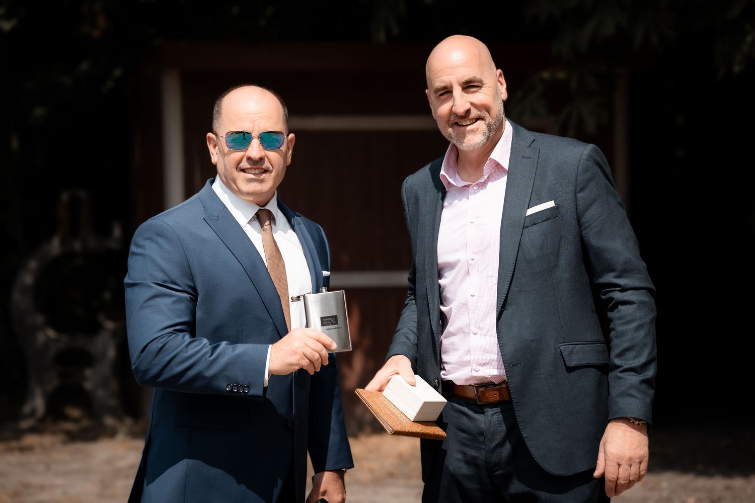 Two men in suits smiling outdoors, one wearing sunglasses holding a gift at a formal event.