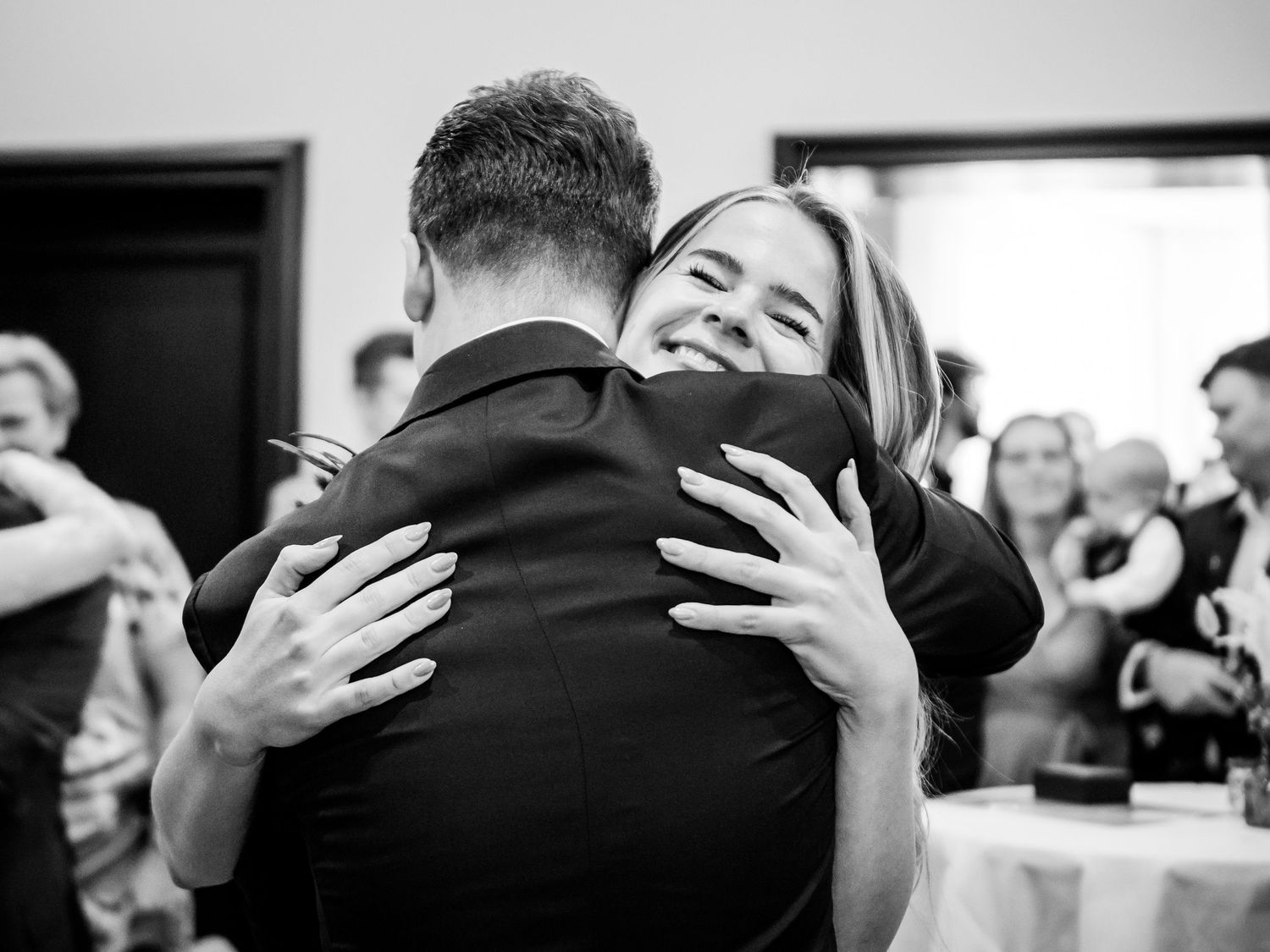 Black and white photo of a woman smiling while sharing a warm embrace at a wedding reception.