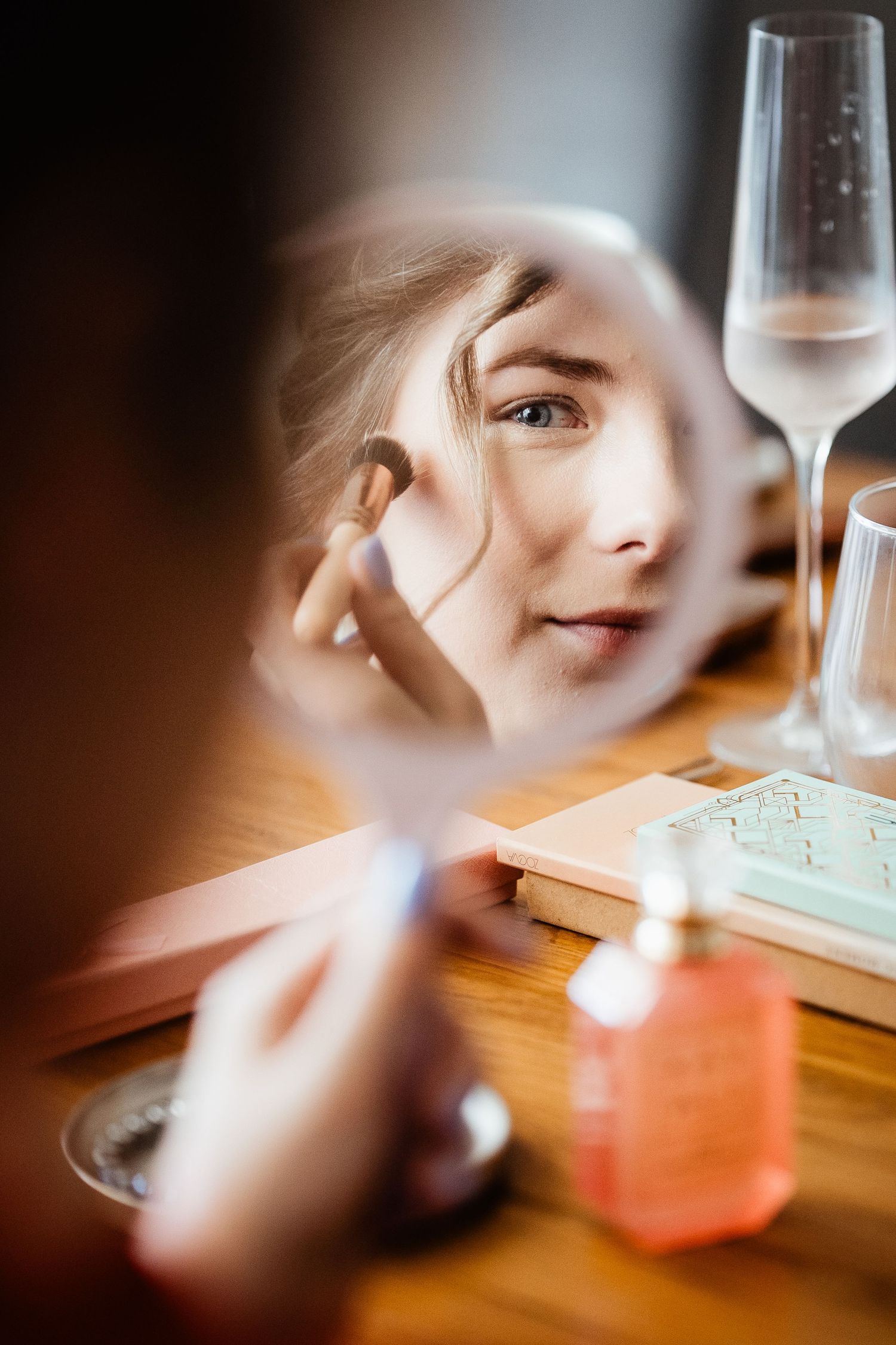 Young woman applying makeup with a brush, reflected in a round mirror on a wooden vanity with beauty products.