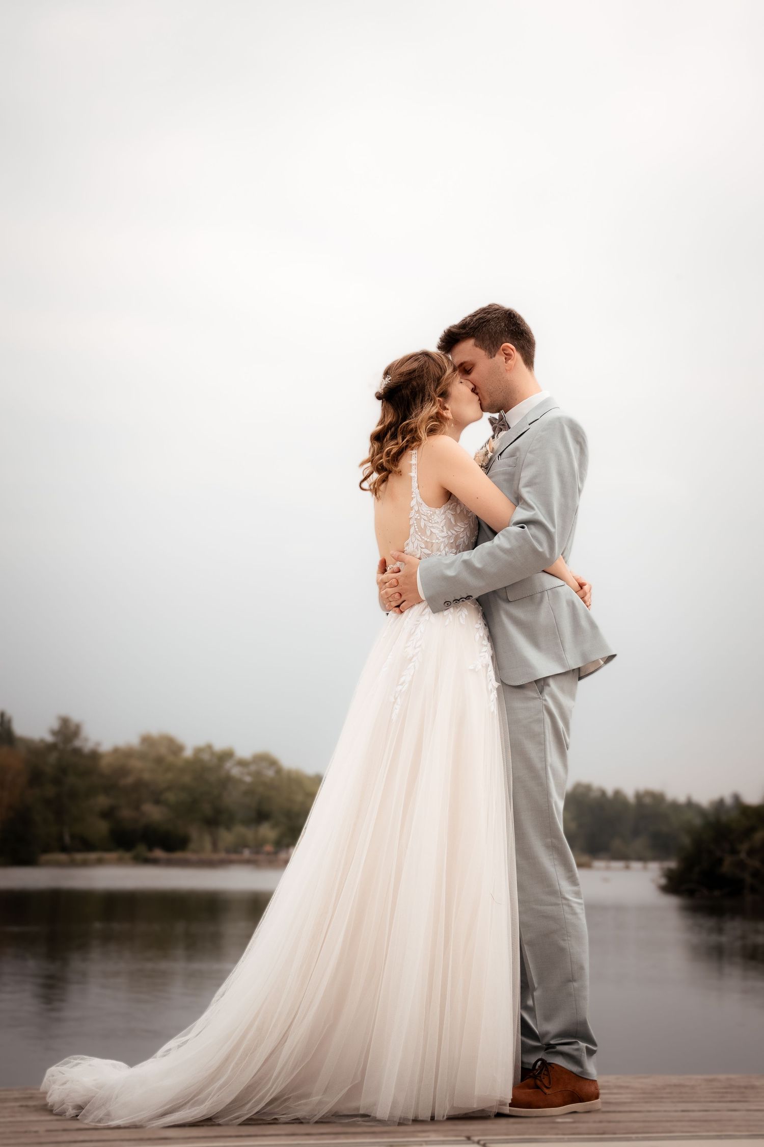 Bride and groom embracing on a wooden dock by a lake, wearing a white gown and gray suit on a cloudy day.
