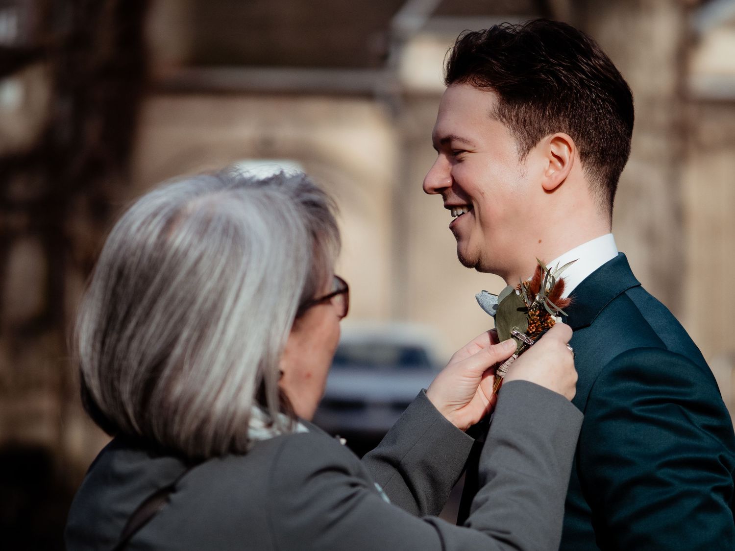 An older woman pins a boutonniere on a smiling groom in a teal suit outdoors before a wedding ceremony.
