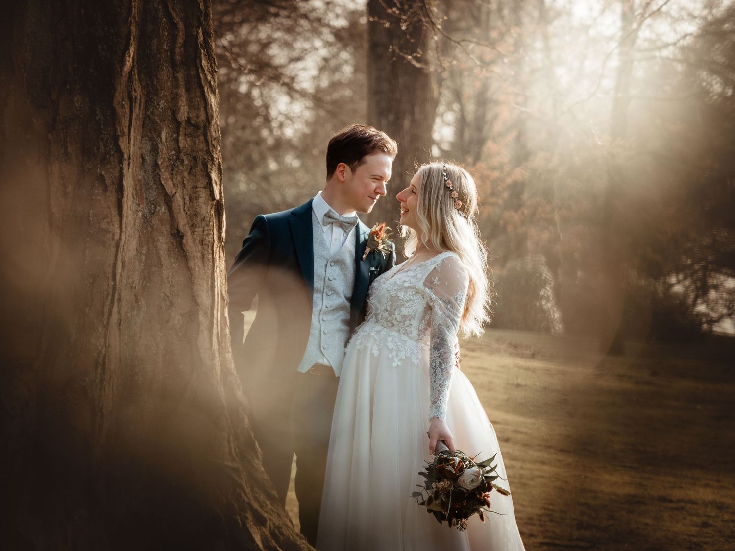 Bride and groom share a romantic moment near a sunlit tree, bride holding a bouquet in an elegant lace wedding gown.