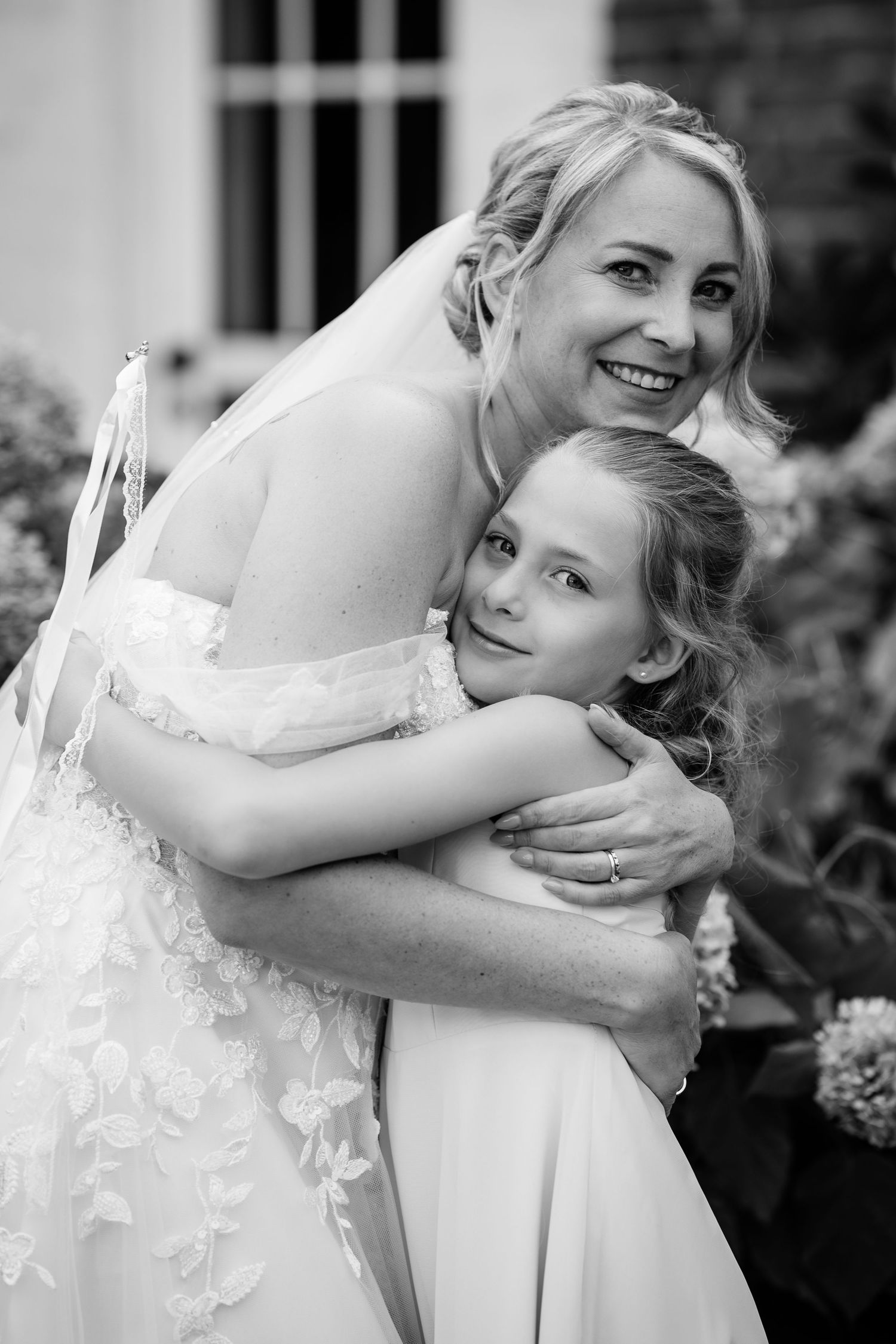 Bride in lace off-shoulder gown embracing a young girl in a black and white wedding portrait.