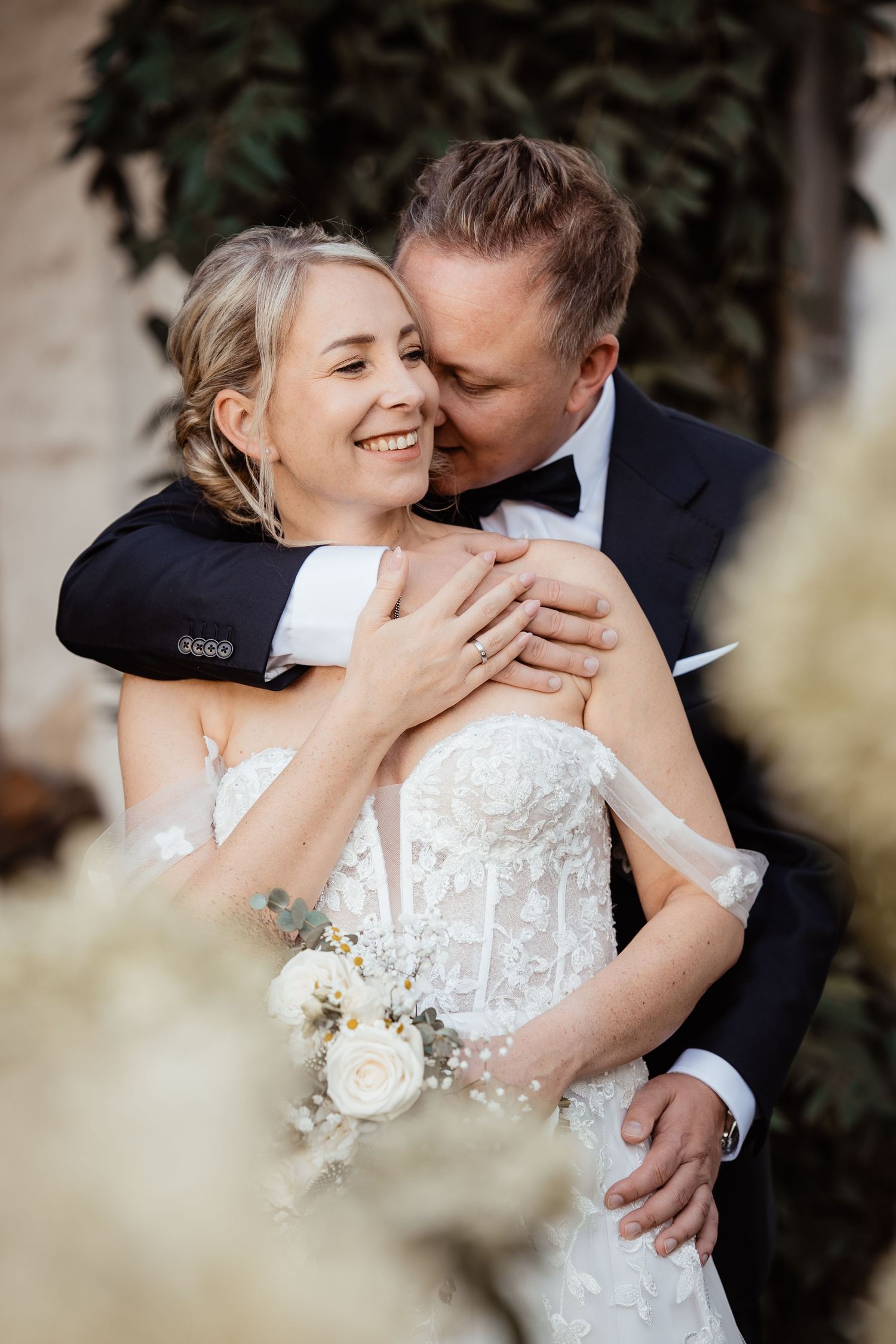 Groom in black tuxedo embracing smiling bride in lace gown with white bouquet.