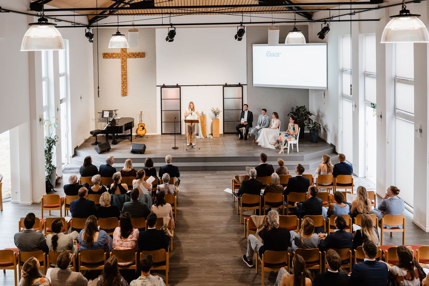 Congregation gathered in a bright modern church with wooden cross during a wedding ceremony.