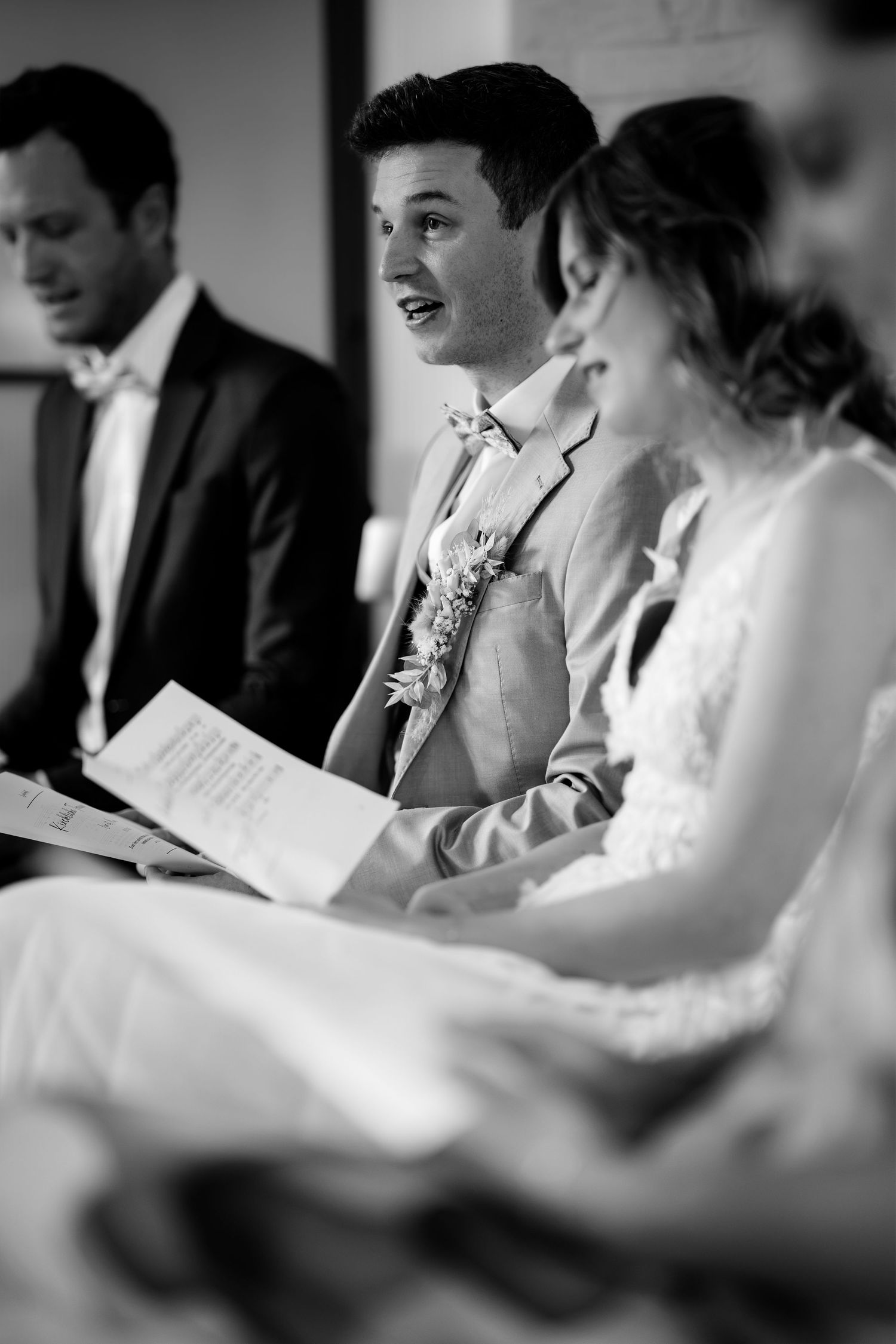 Groom in a light suit reading vows beside his bride during a wedding ceremony in black and white.