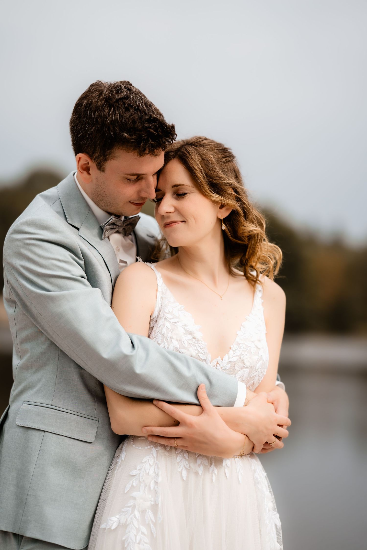 Romantic couple embracing tenderly by a lake, groom in grey suit with bride in lace gown.