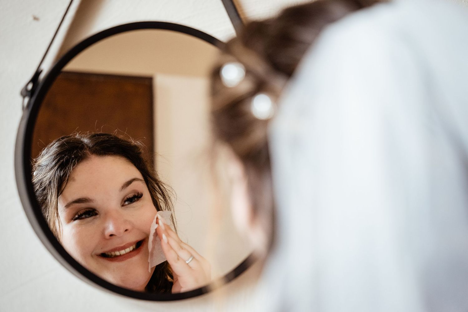 Smiling bride applies makeup while looking into a round mirror during wedding morning preparations.