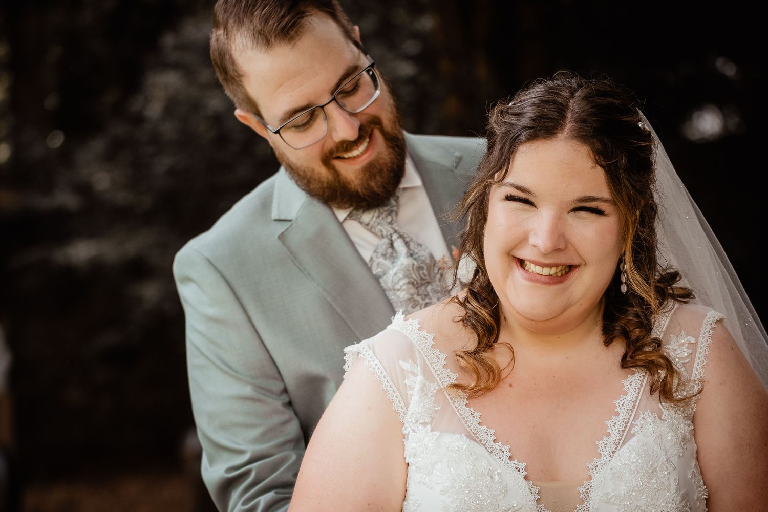 Smiling bride in lace gown and veil with groom in gray suit laughing together during outdoor wedding portrait.