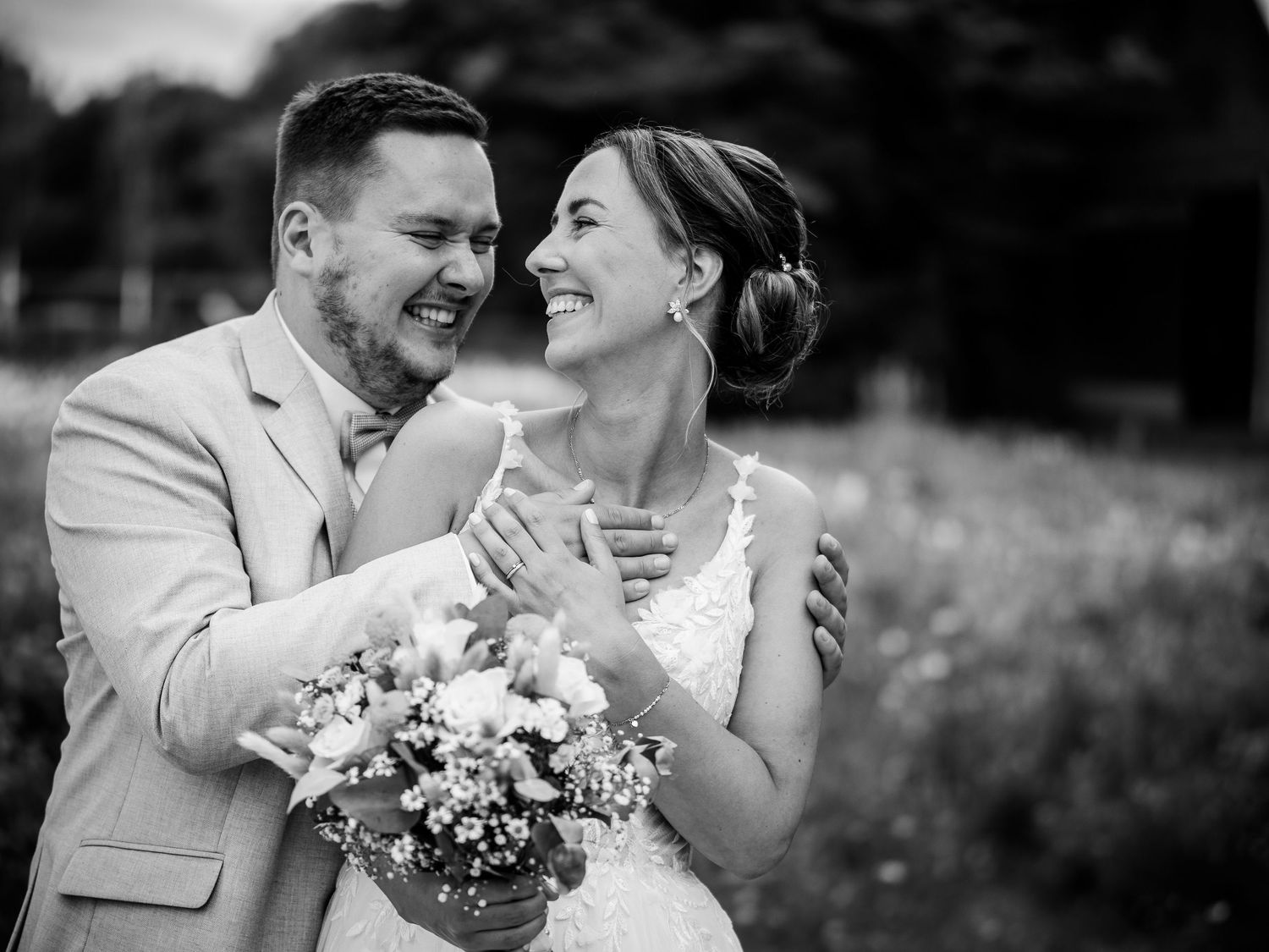 A joyful black and white wedding photo of a couple sharing a candid laughing moment while holding a floral bouquet.