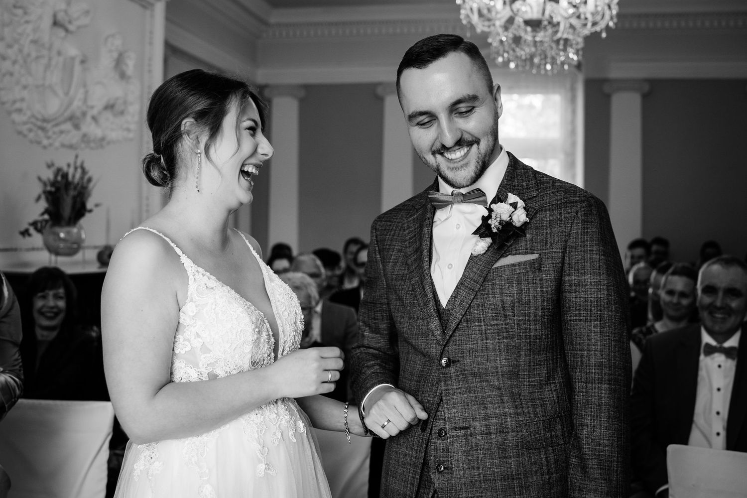 Black and white photo of a laughing bride in lace gown and groom in plaid suit holding hands at wedding ceremony.