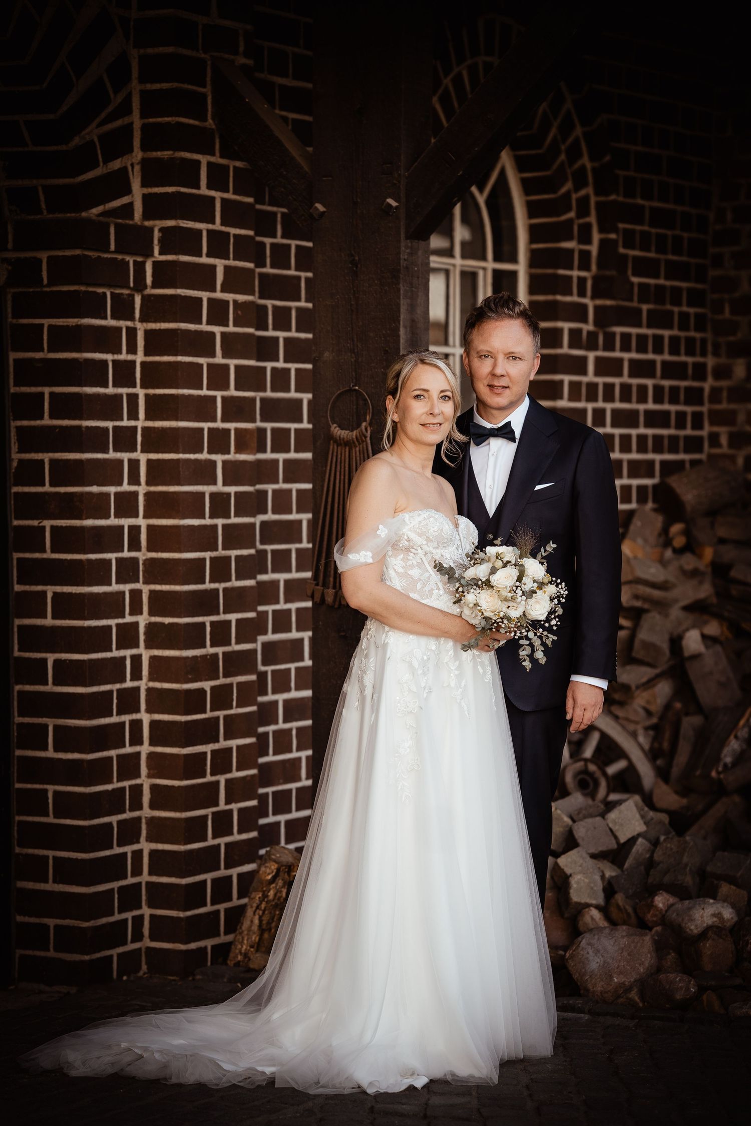 Bride and groom posing in front of a rustic brick wall, bride holding a white bouquet in a strapless gown.