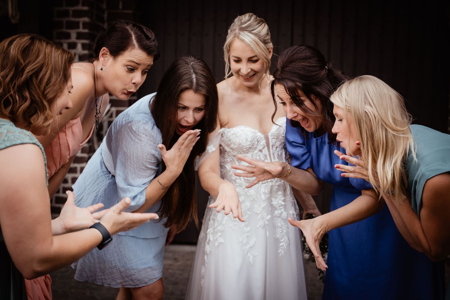 Bride in lace wedding gown surrounded by admiring bridesmaids in colorful dresses.