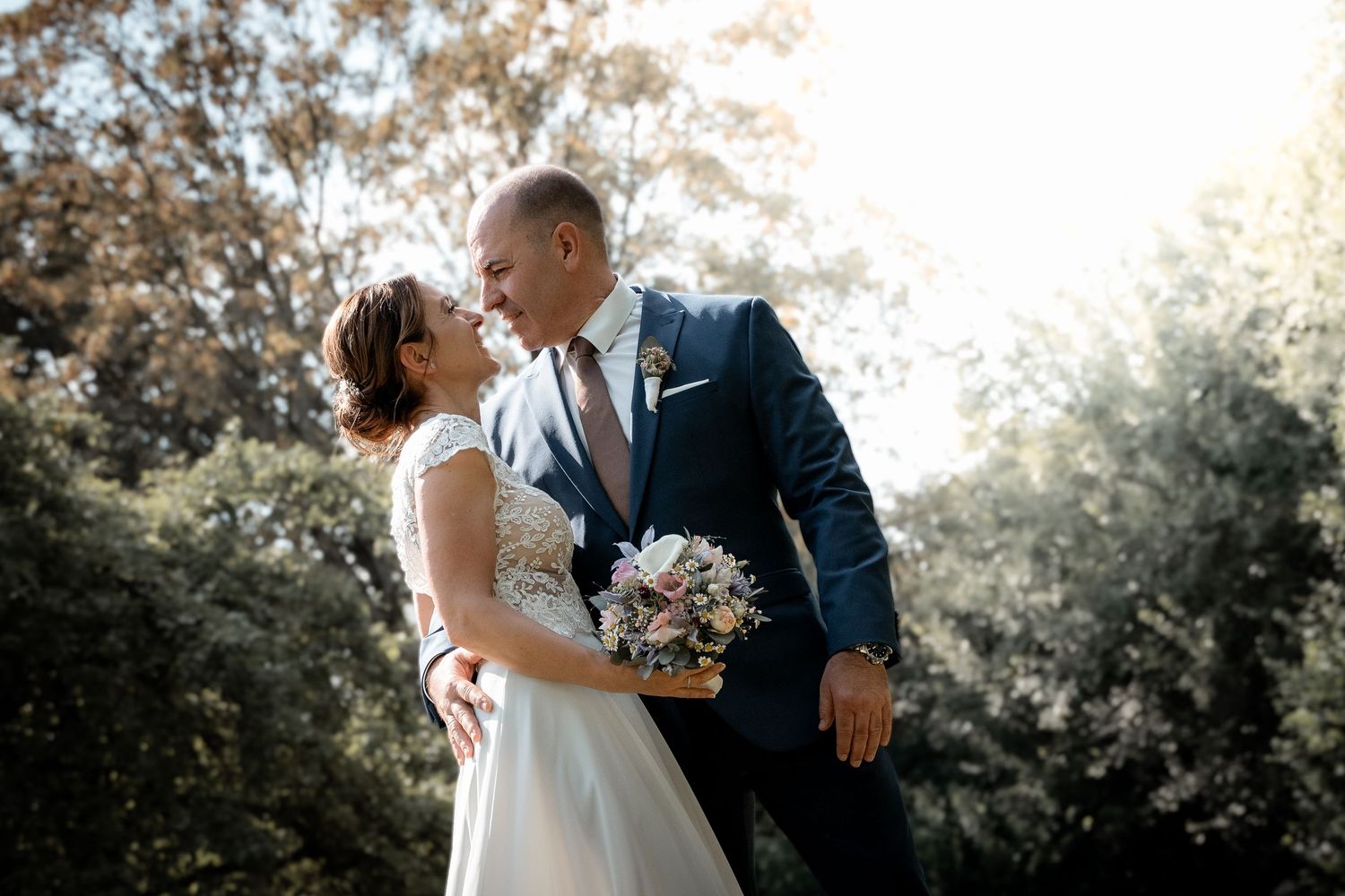 Bride and groom share a romantic moment outdoors, surrounded by lush green trees on their wedding day.