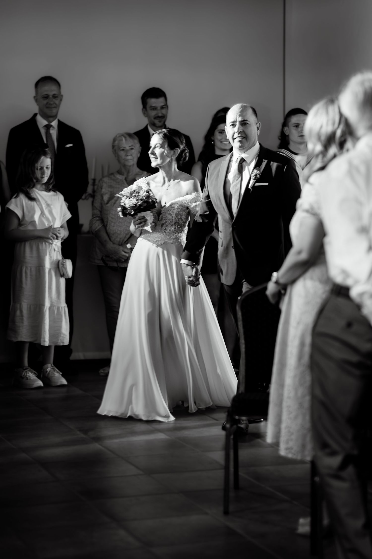 Bride walks down the aisle with escort during black and white wedding ceremony with guests watching.