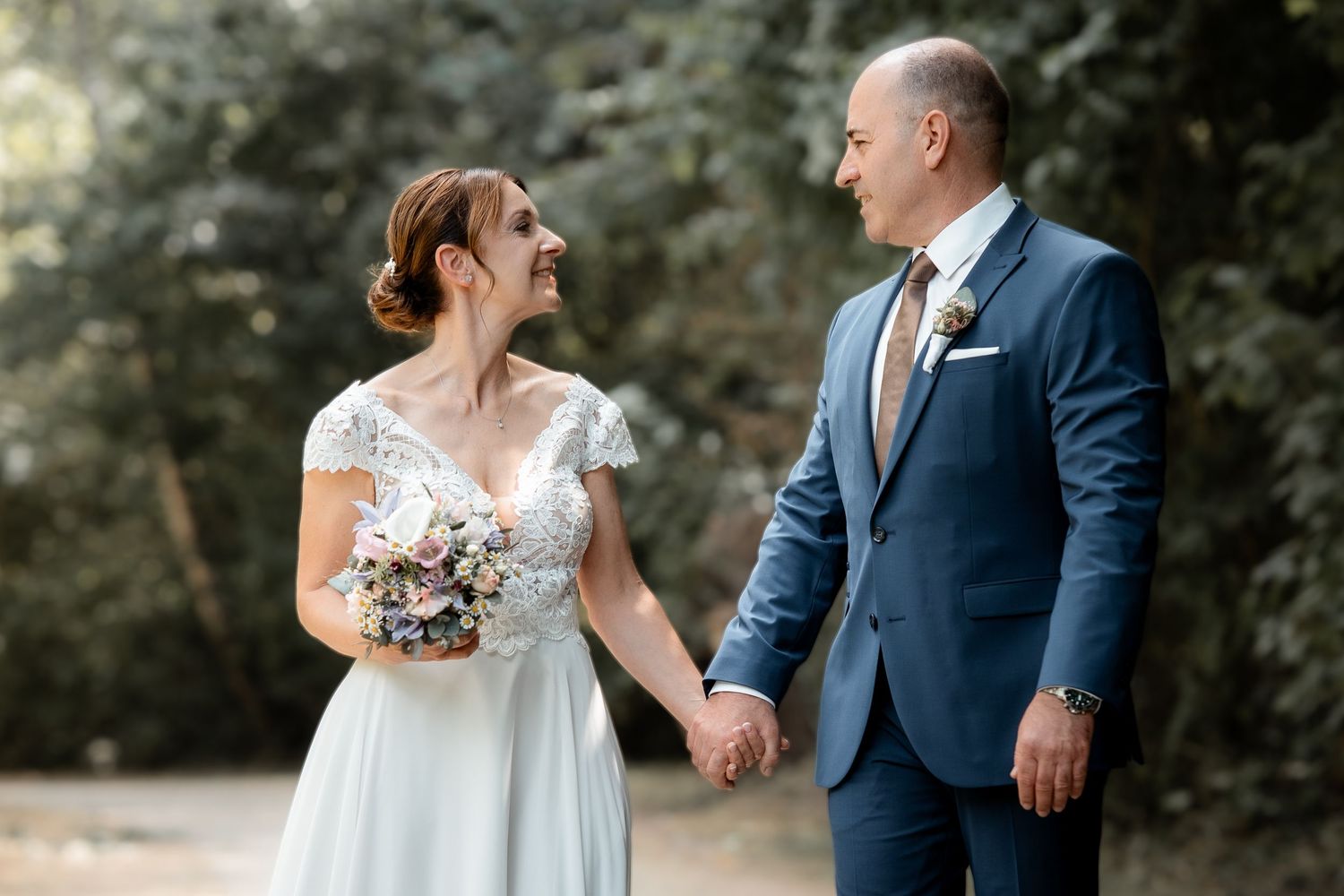 Bride and groom holding hands smiling at each other outdoors, bride in lace dress holding bouquet, groom in blue suit.