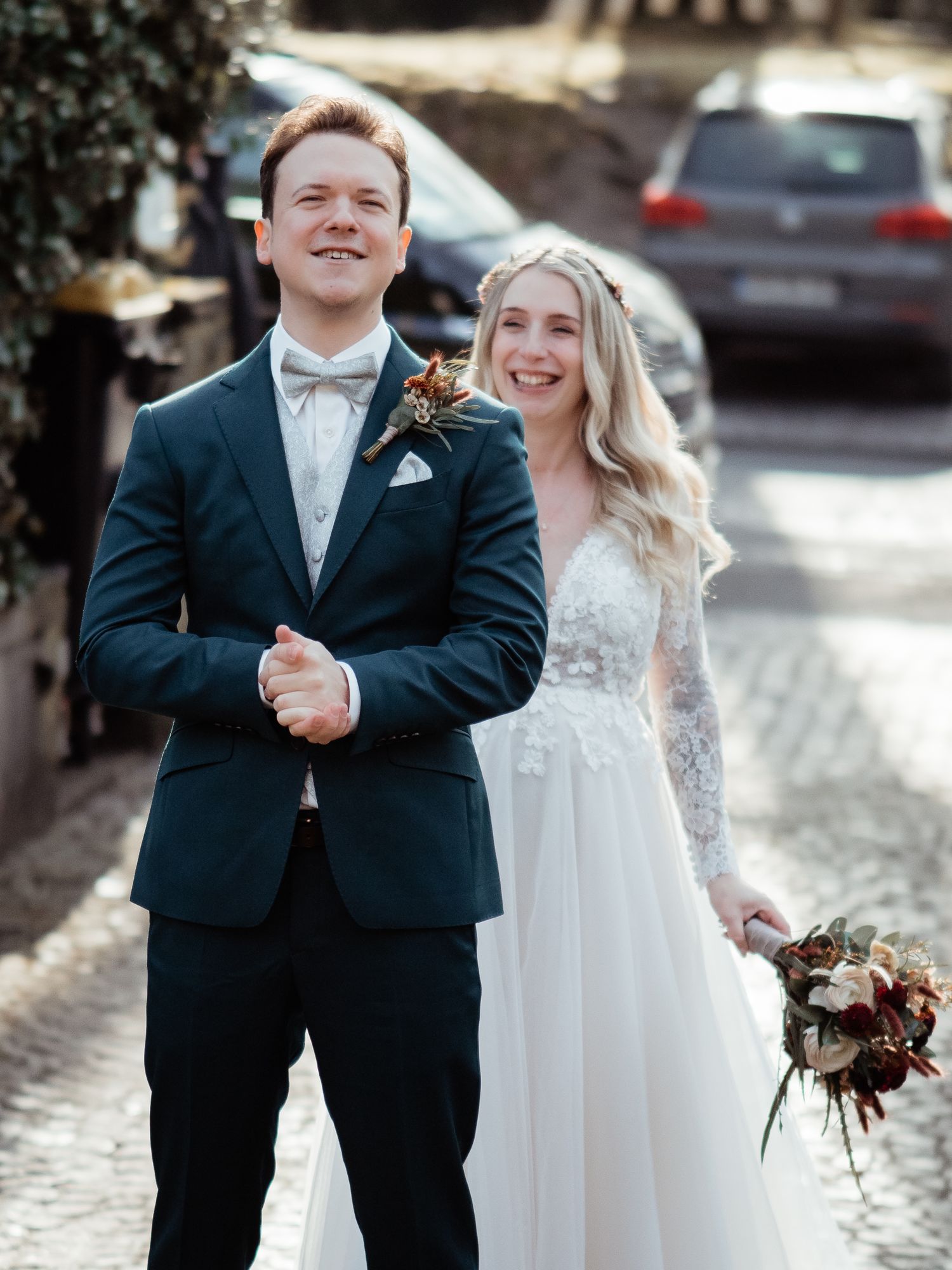 Smiling bride and groom walking together outdoors, bride holding a bouquet in a lace wedding gown.