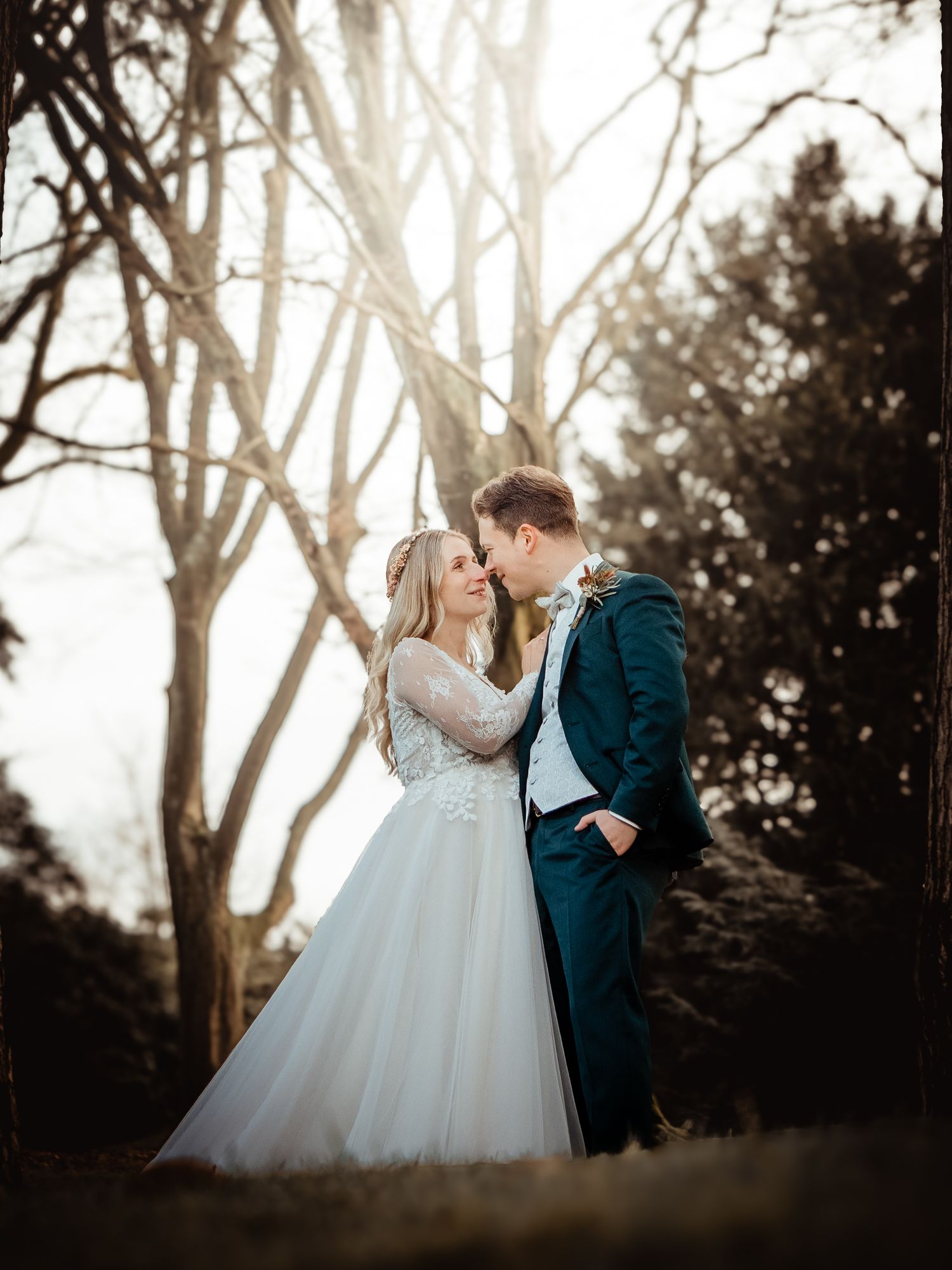 A bride and groom share a romantic moment outdoors, surrounded by bare trees with golden sunlight glowing behind them.