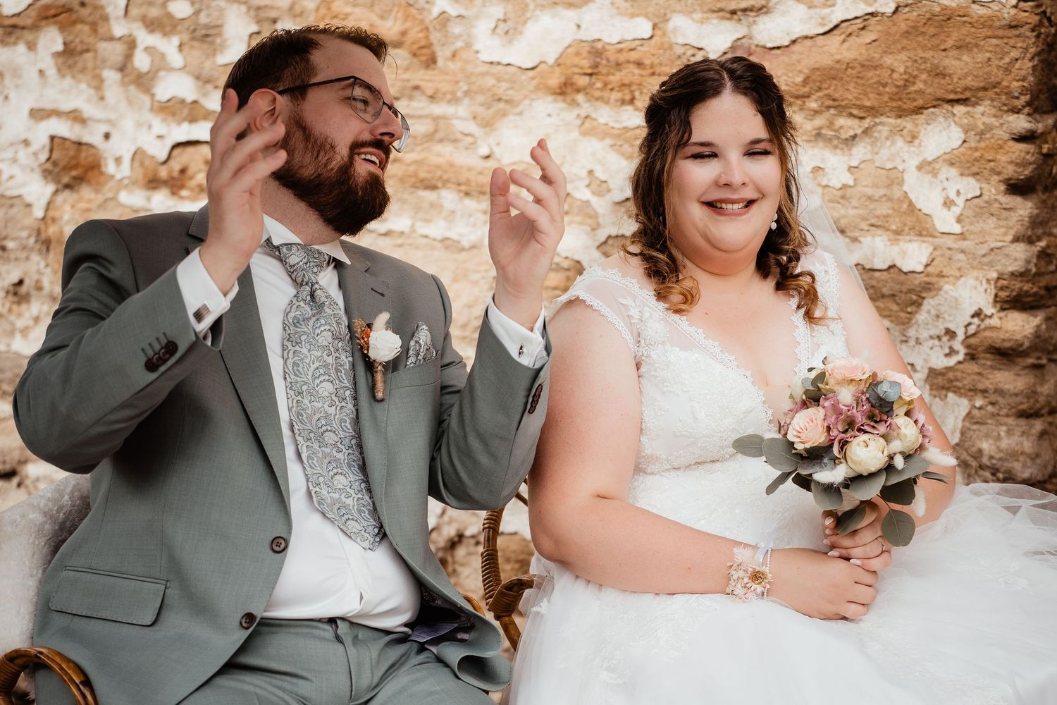 Laughing bride holding bouquet and groom in gray suit sitting together at rustic stone wall wedding venue.