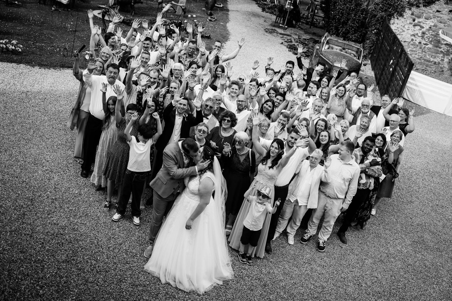 Aerial black and white wedding photo of happy couple kissing surrounded by cheering guests.
