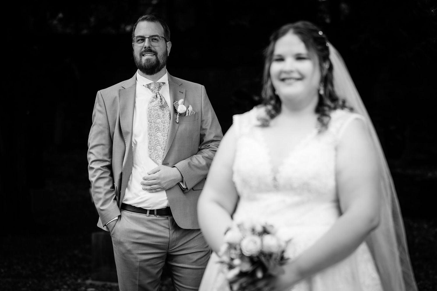 Groom in gray suit watches bride holding bouquet in black and white wedding photo outdoors.