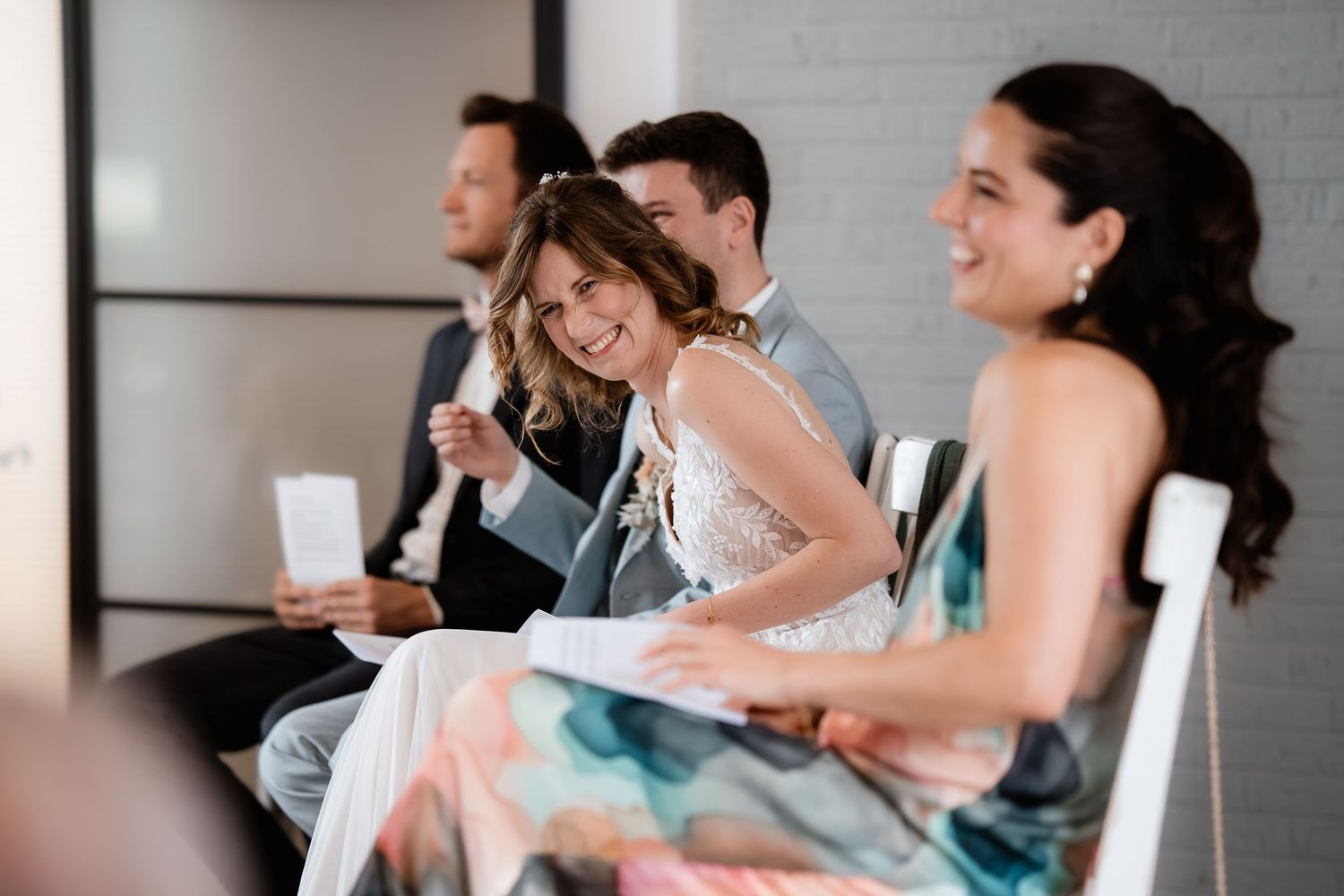 A bride laughs joyfully with guests during a wedding ceremony seated in white chairs.