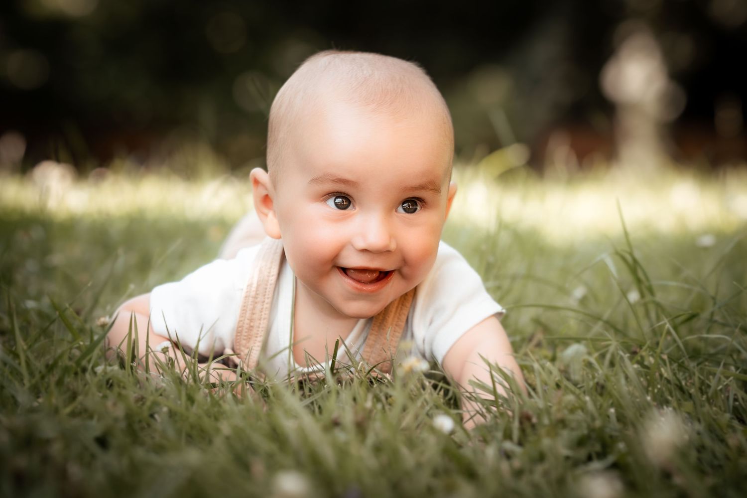Smiling baby lying on green grass outdoors, wearing a white onesie, with a blurred natural background.