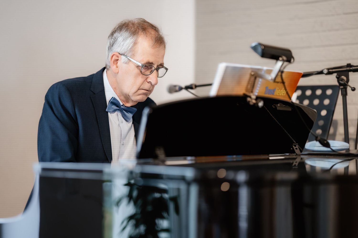 Elderly pianist in suit and bow tie playing grand piano while reading sheet music on stand.