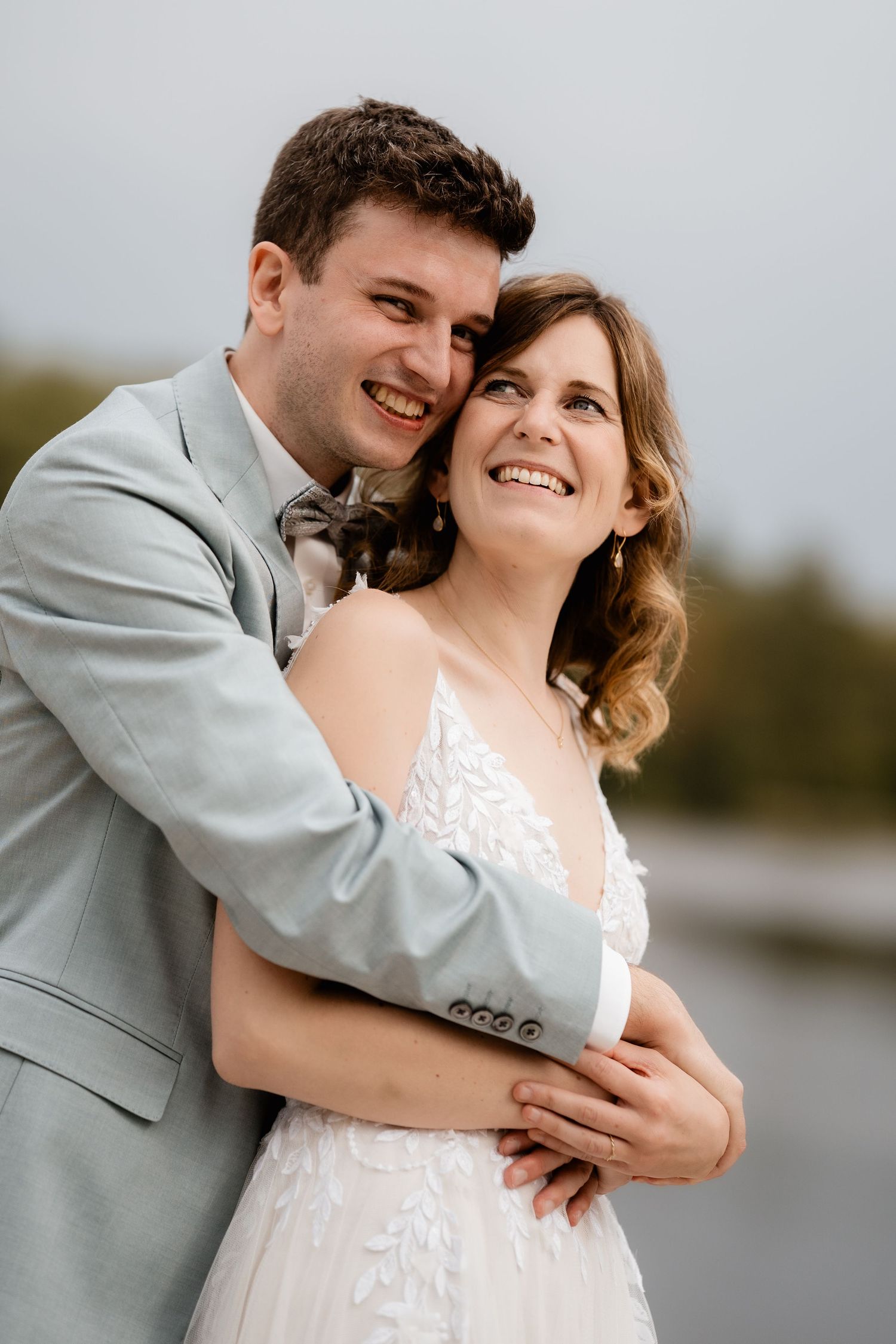 A smiling bride and groom embrace closely outdoors near a lake on their wedding day.