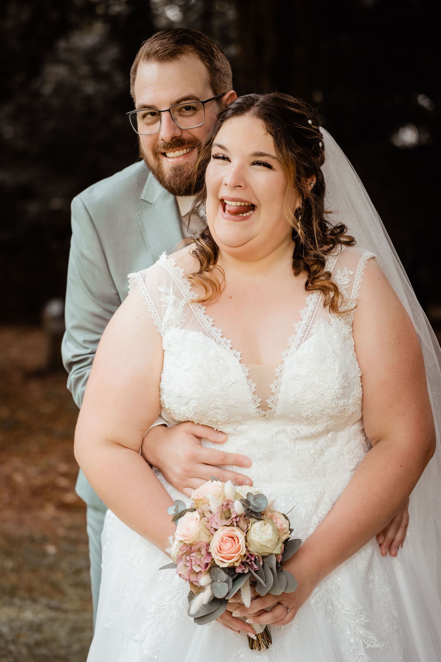 Smiling bride in white lace gown holds bouquet while groom in sage suit embraces her outdoors.