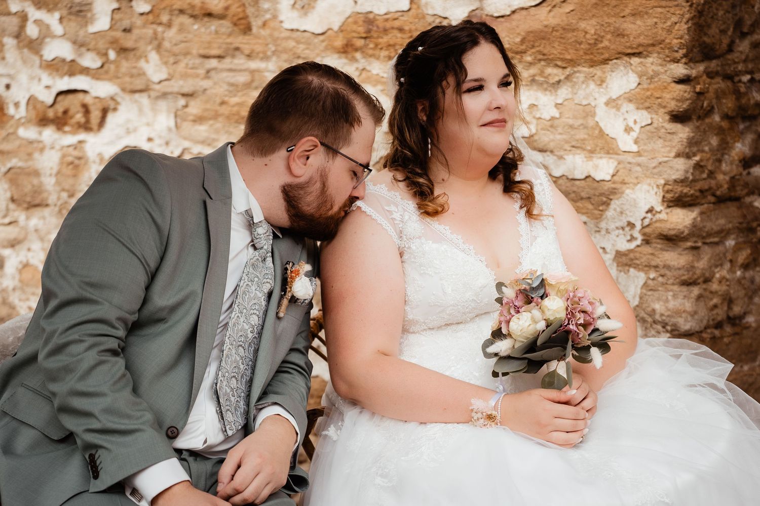 Bride and groom sitting together against rustic stone wall, bride holding bouquet of flowers in wedding portrait.