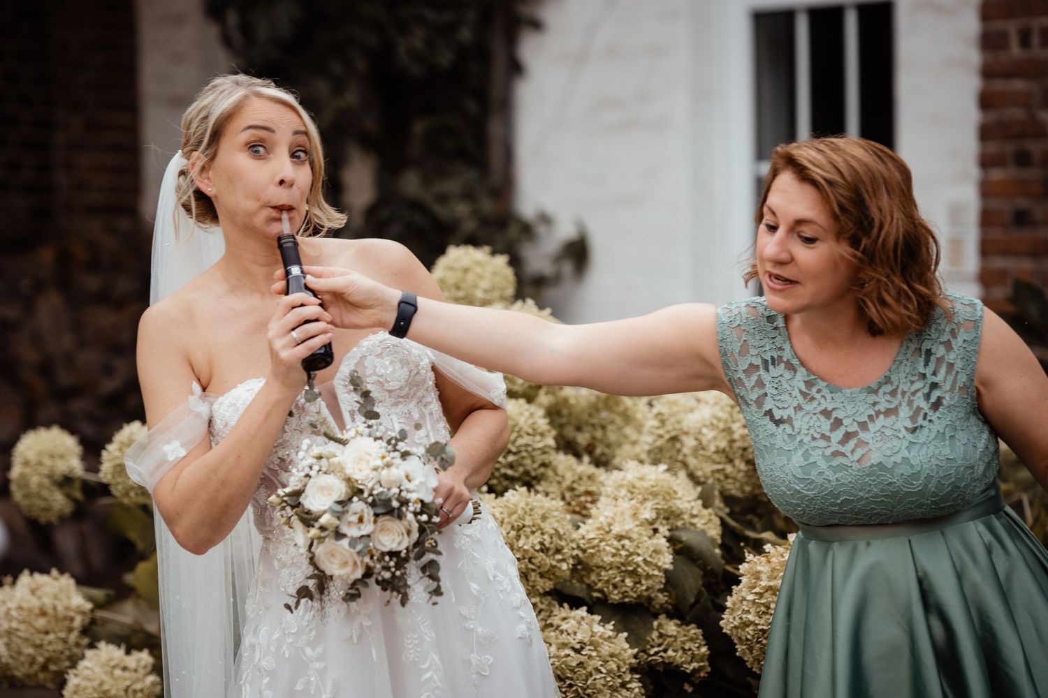 Bride holding bouquet drinks from a flask assisted by bridesmaid in teal dress outdoors.