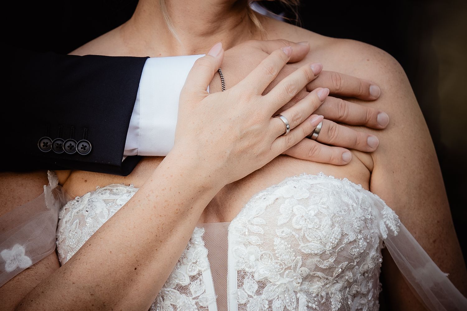 Close-up of bride and groom hands on wedding day, showcasing diamond engagement ring on lace bridal gown.