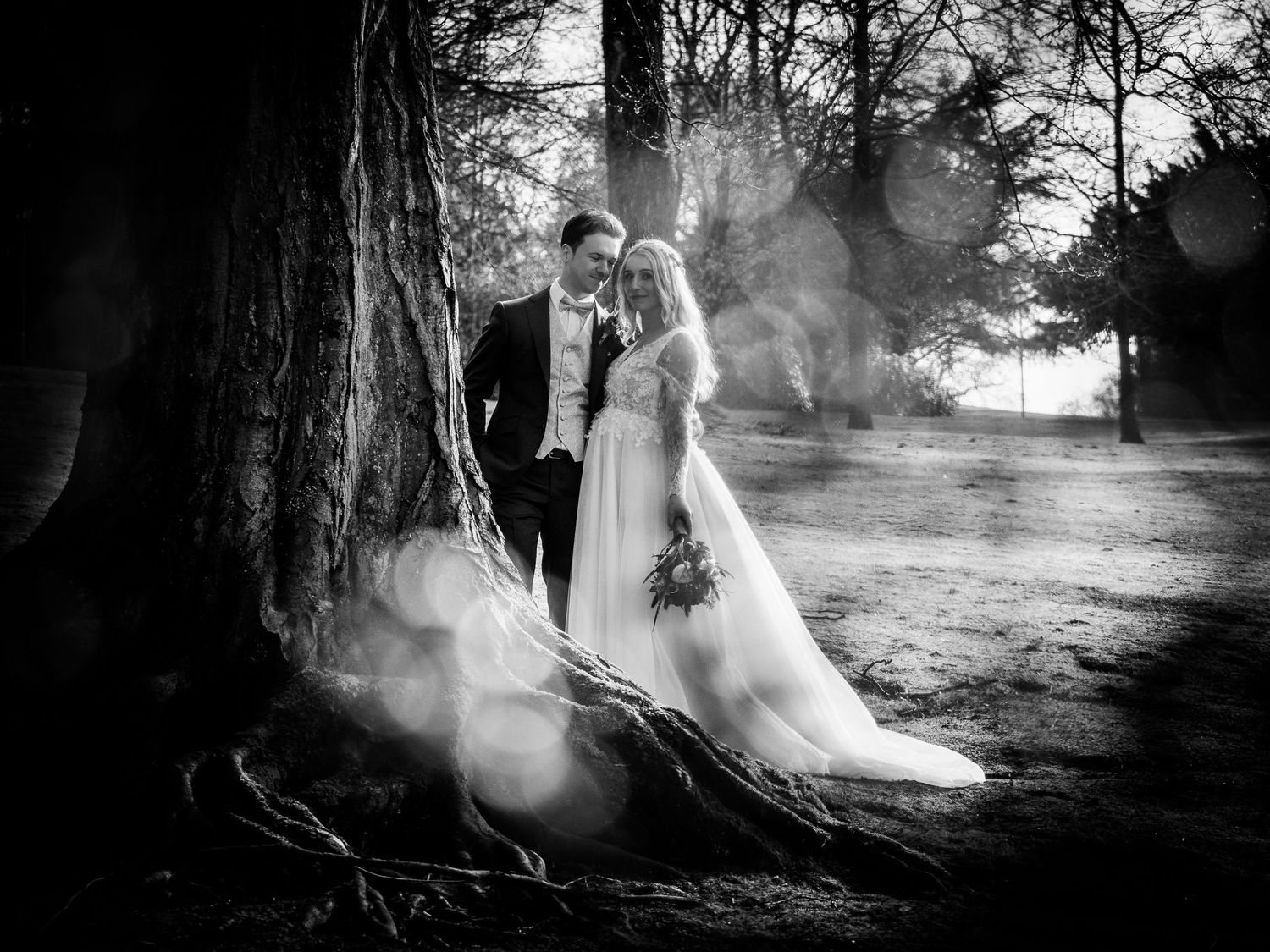 Black and white romantic portrait of bride and groom standing beneath a large tree with sunlight.