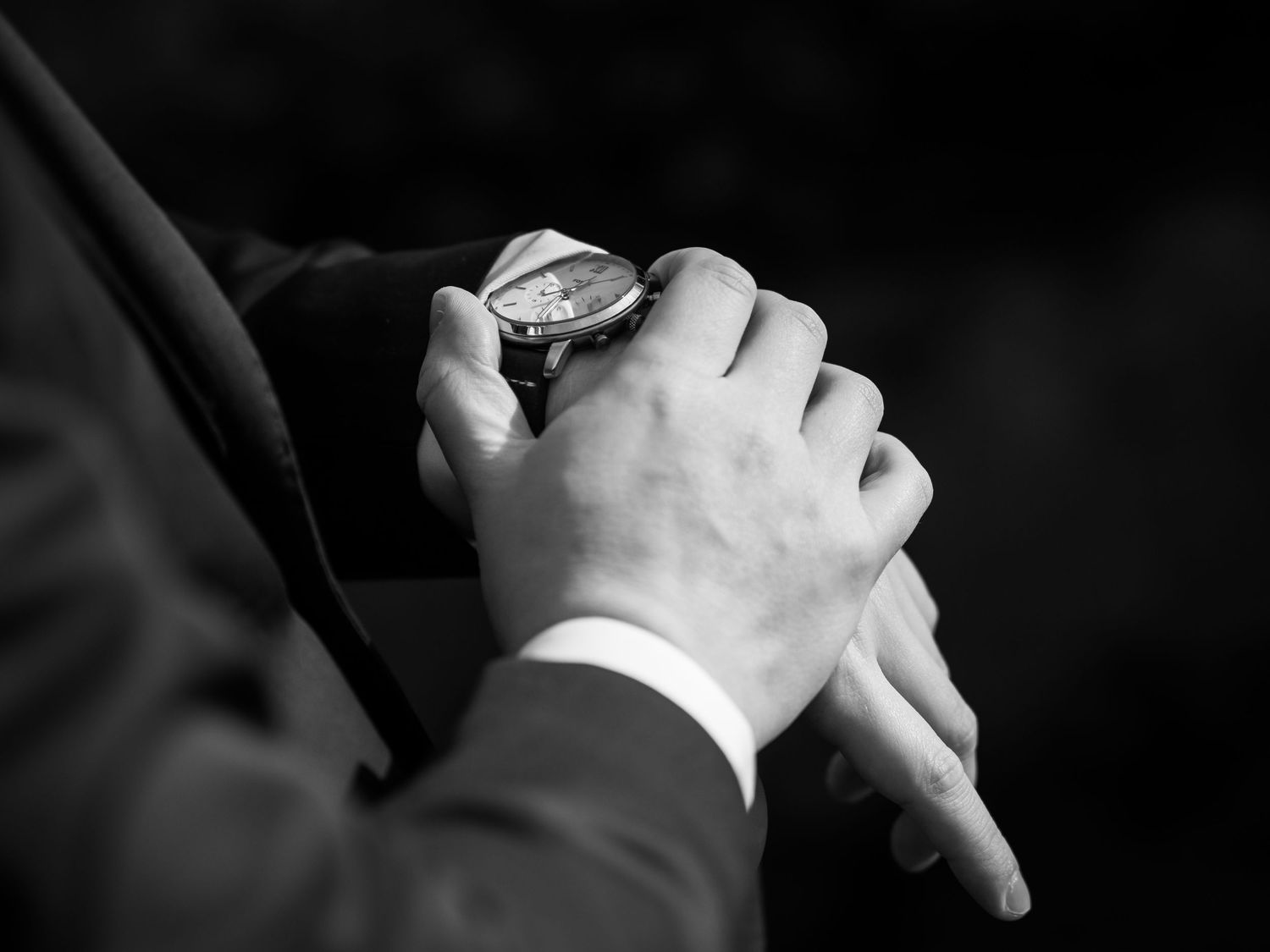 Black and white photo of a man in a suit checking a luxury wristwatch, clasping his hands together.