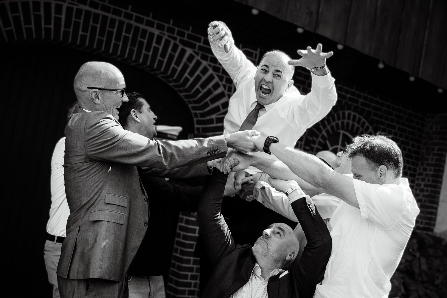 Groomsmen joyfully lifting groom in black and white action shot at outdoor wedding celebration.
