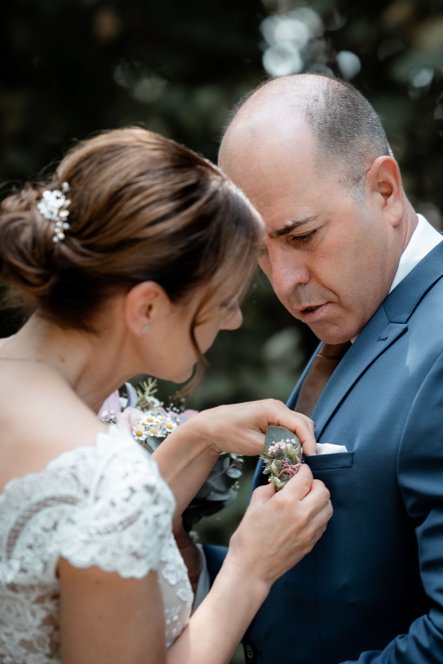 Bride in lace dress pinning boutonniere on groom's navy suit lapel at outdoor wedding ceremony.