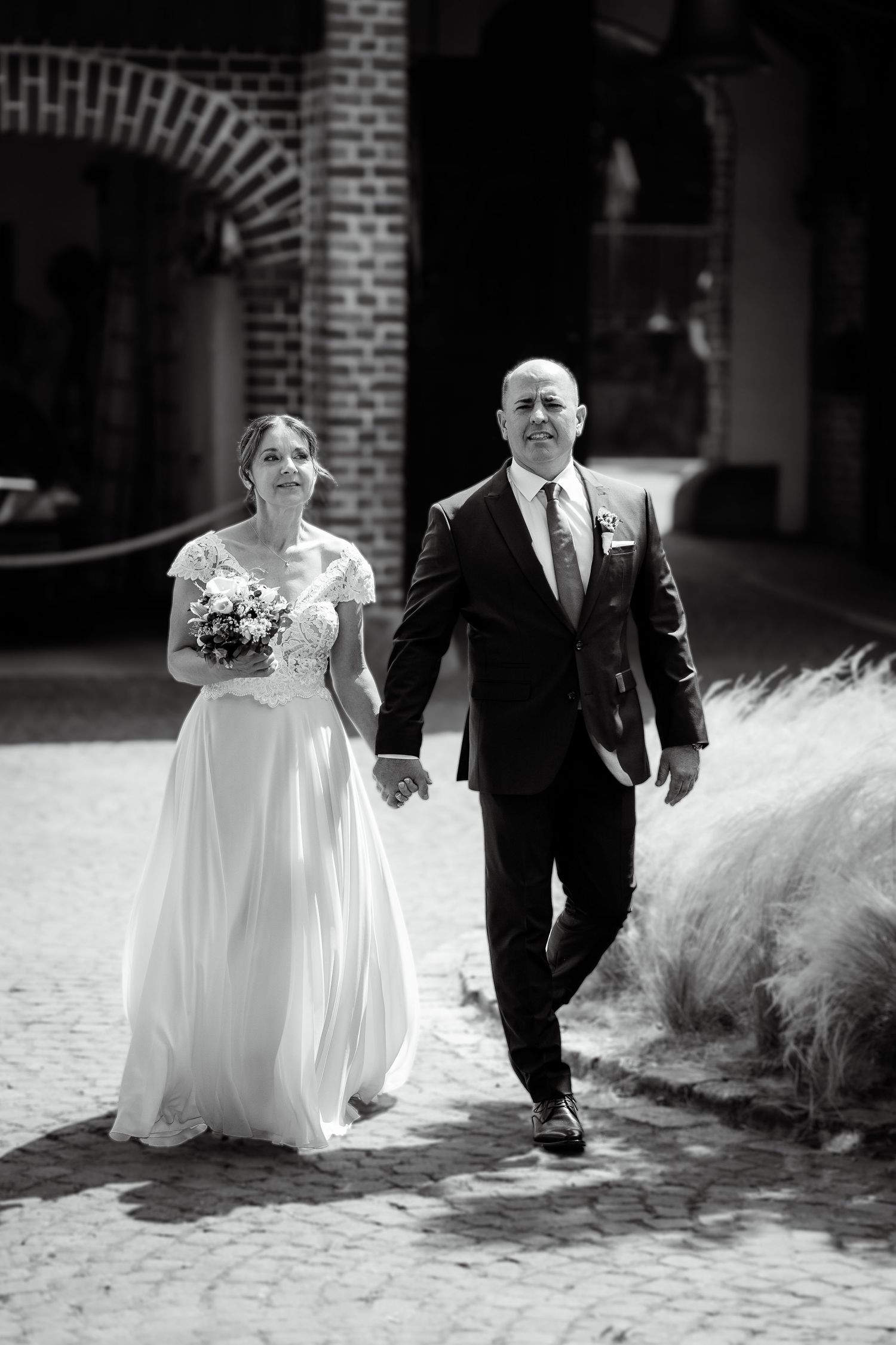 Black and white photo of a bride and groom holding hands walking together on cobblestone path.