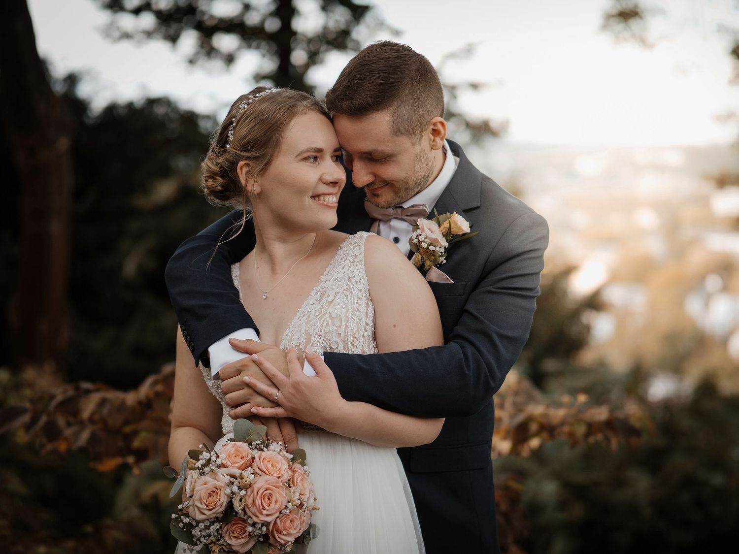 Romantic wedding couple embraces outdoors during golden hour with peach roses in a dreamy forest setting.